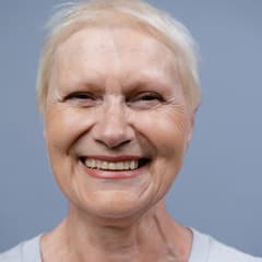 Portrait carré d'une femme âgée aux cheveux blancs courts, souriante sur fond bleu-gris