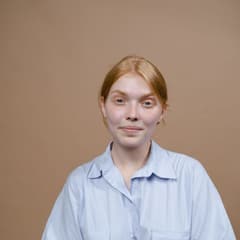 Portrait carré d'une jeune femme rousse en chemise bleu clair, expression neutre sur fond beige