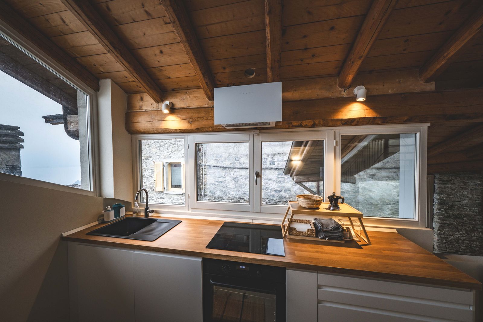 Cuisine ouverte au plafond à poutres de bois, plan de travail bois et vue sur façade en pierre