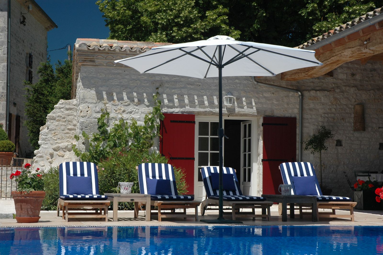 Façade de mas en pierre claire aux volets rouges, transats rayés bleus au bord de la piscine