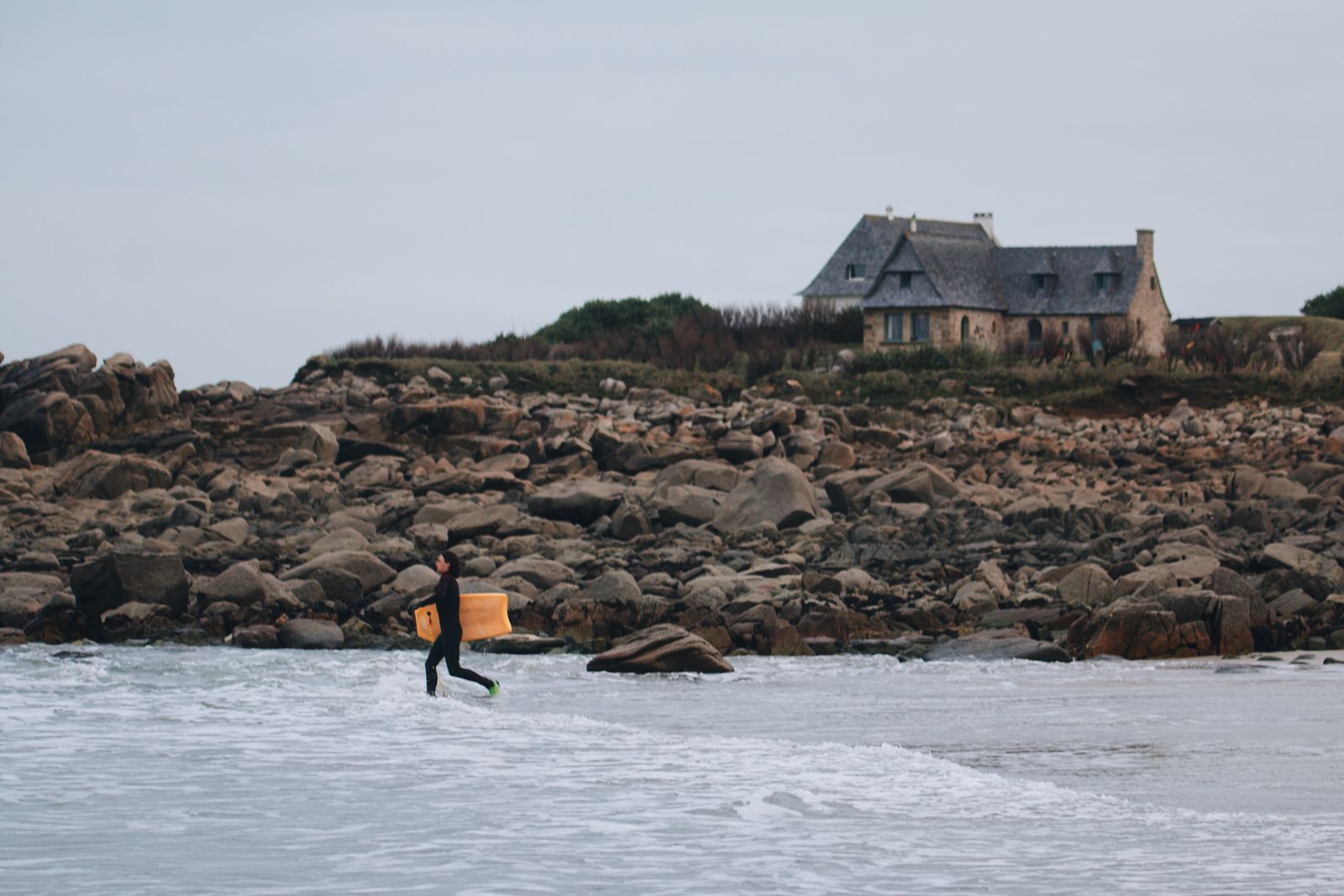 Surfeur en combinaison courant vers les vagues, maison de granit sur les rochers en arrière-plan