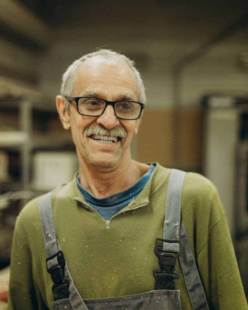 Artisan souriant à lunettes et moustache, en pull vert et cotte à bretelles, dans son atelier