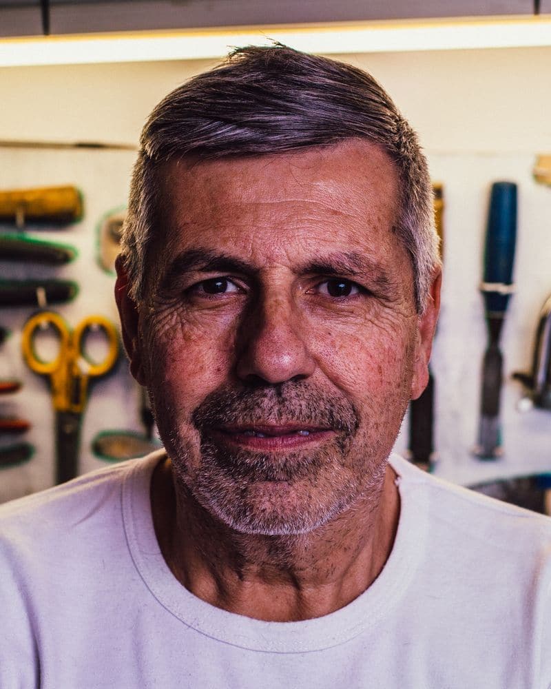 Portrait d'un artisan d'une soixantaine d'années en tee-shirt clair, devant un panneau d'outils d'atelier