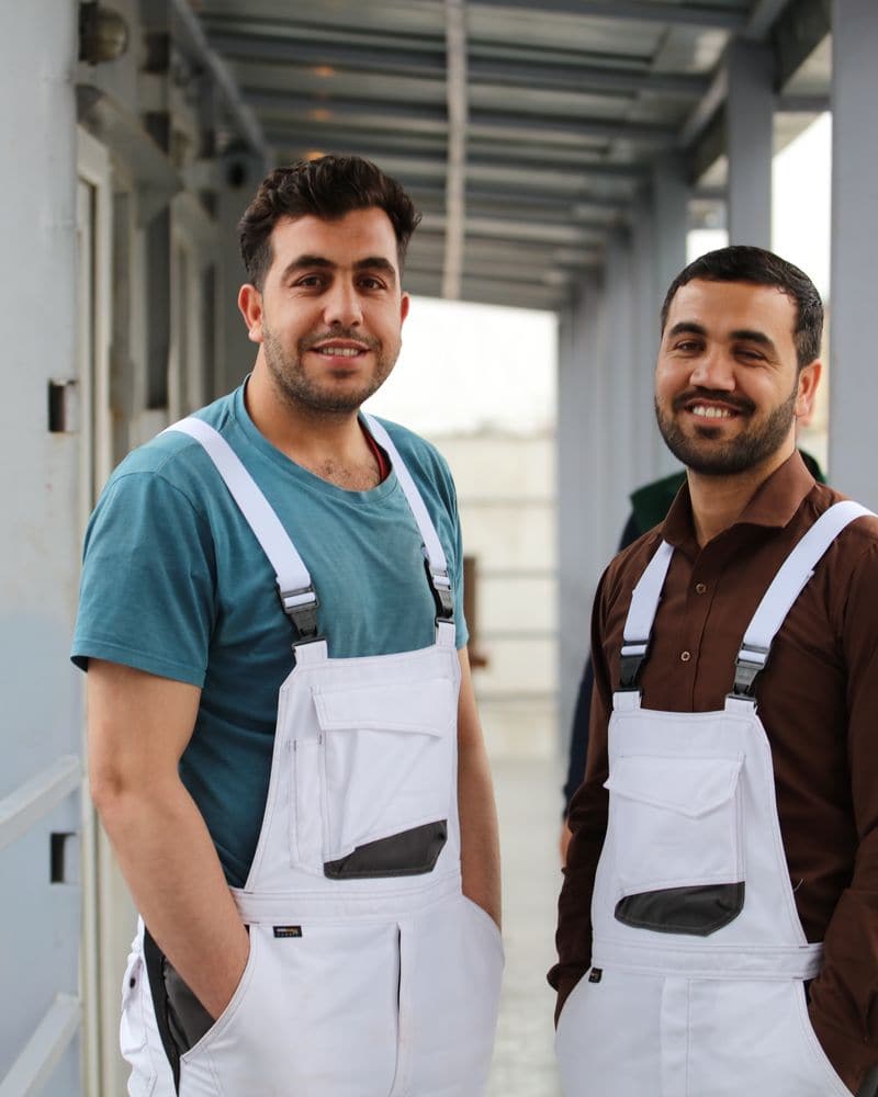 Binôme de deux artisans souriants en cottes à bretelles blanches, dans la coursive d'un bâtiment