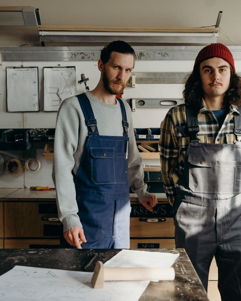 Duo d'artisans de deux générations en cottes à bretelles, debout derrière un établi dans leur atelier
