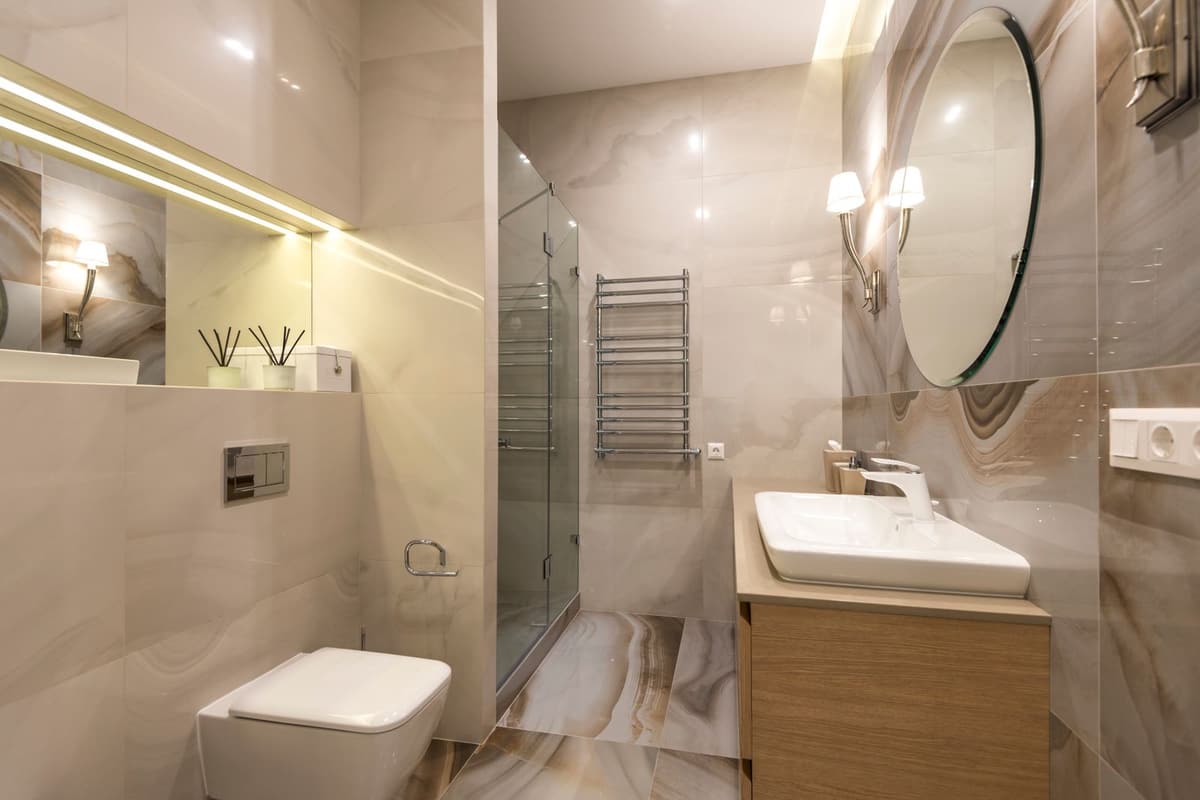 Salle de bain habillée de marbre beige avec grand miroir rond, meuble bois et douche vitrée